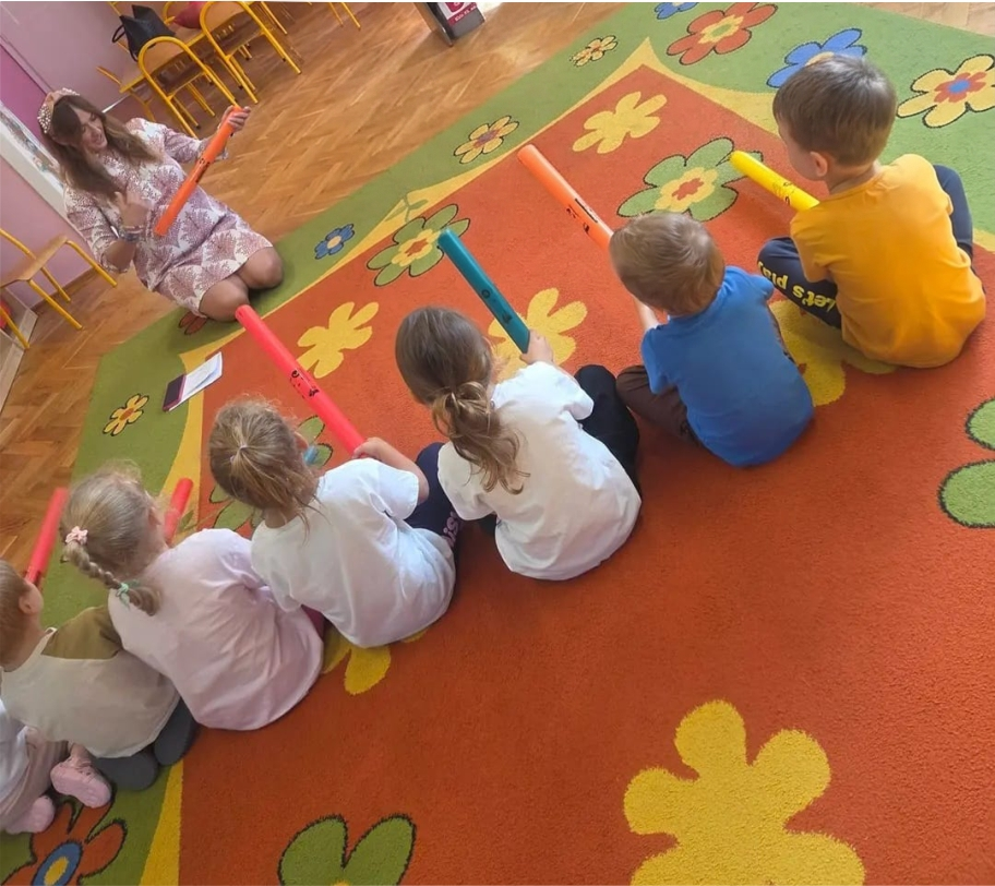 Dzieci siedzą same na dywanie z kolorowymi Bum Bum Rurkami i patrzą na Joannę Stachowską podczas zajęć rytmiki